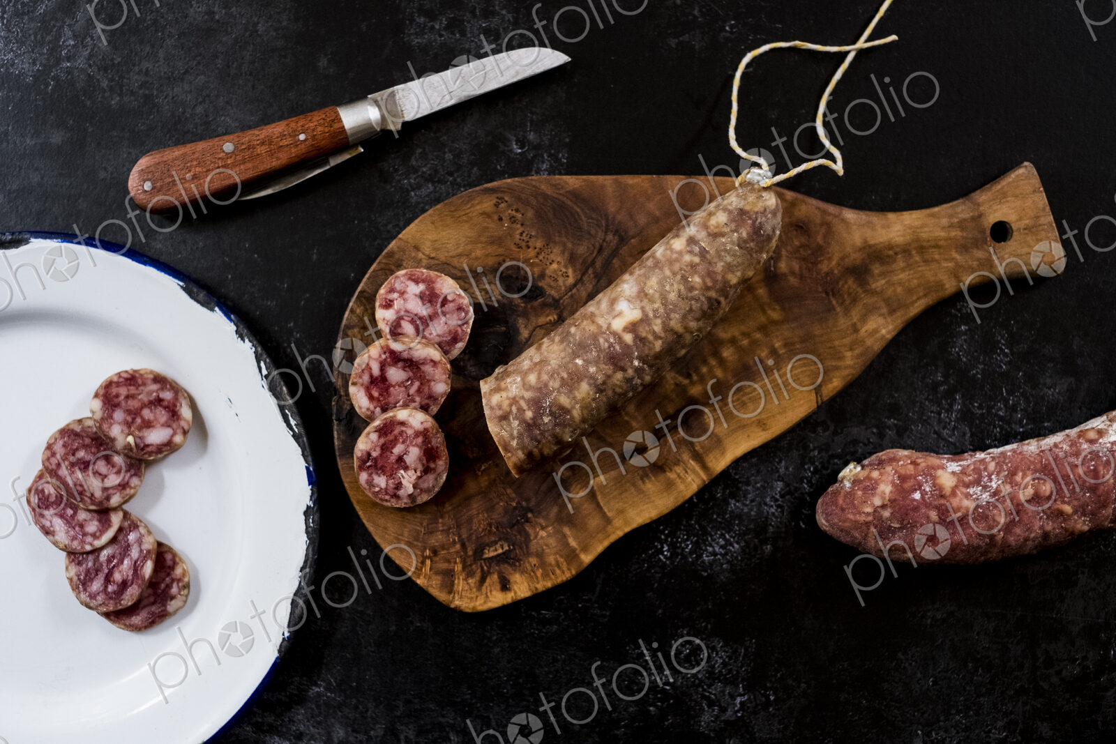 High angle close up of knife, sliced salami on wooden cutting board and white enamel…