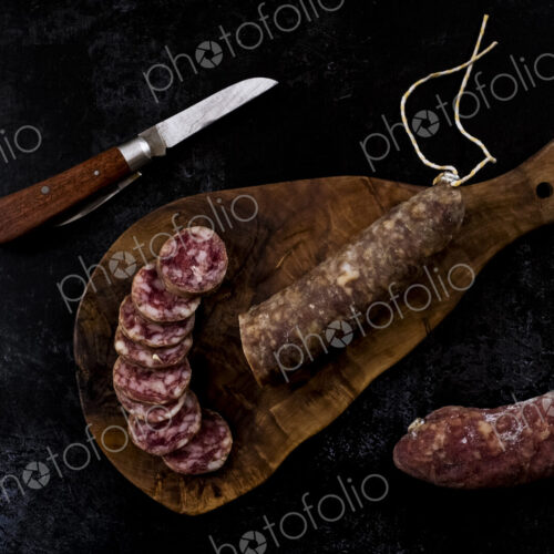 High angle close up of knife, sliced salami on wooden cutting board on black background. High angle close up of knife, sliced salami on wooden cutting board on black background.