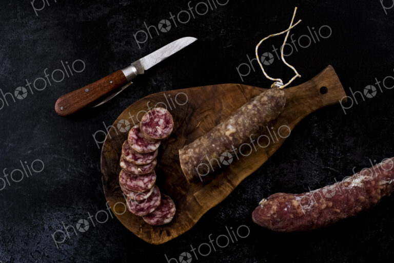 High angle close up of knife, sliced salami on wooden cutting board on black background.