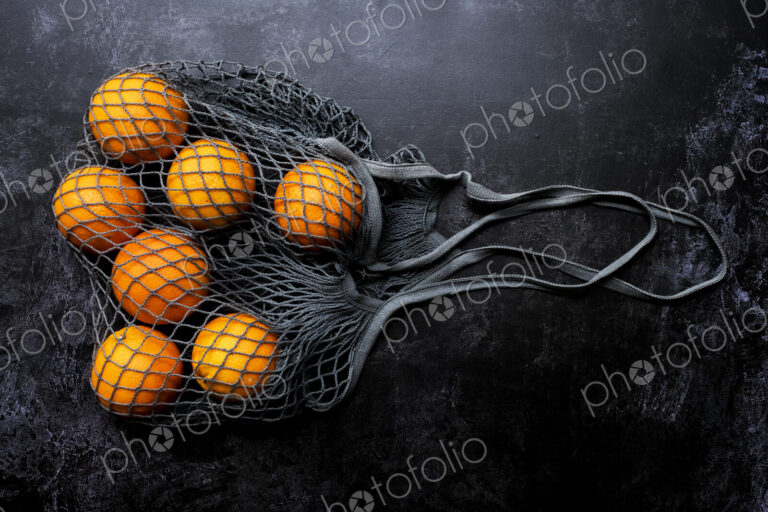 High angle close up of oranges in grey net bag on black background.