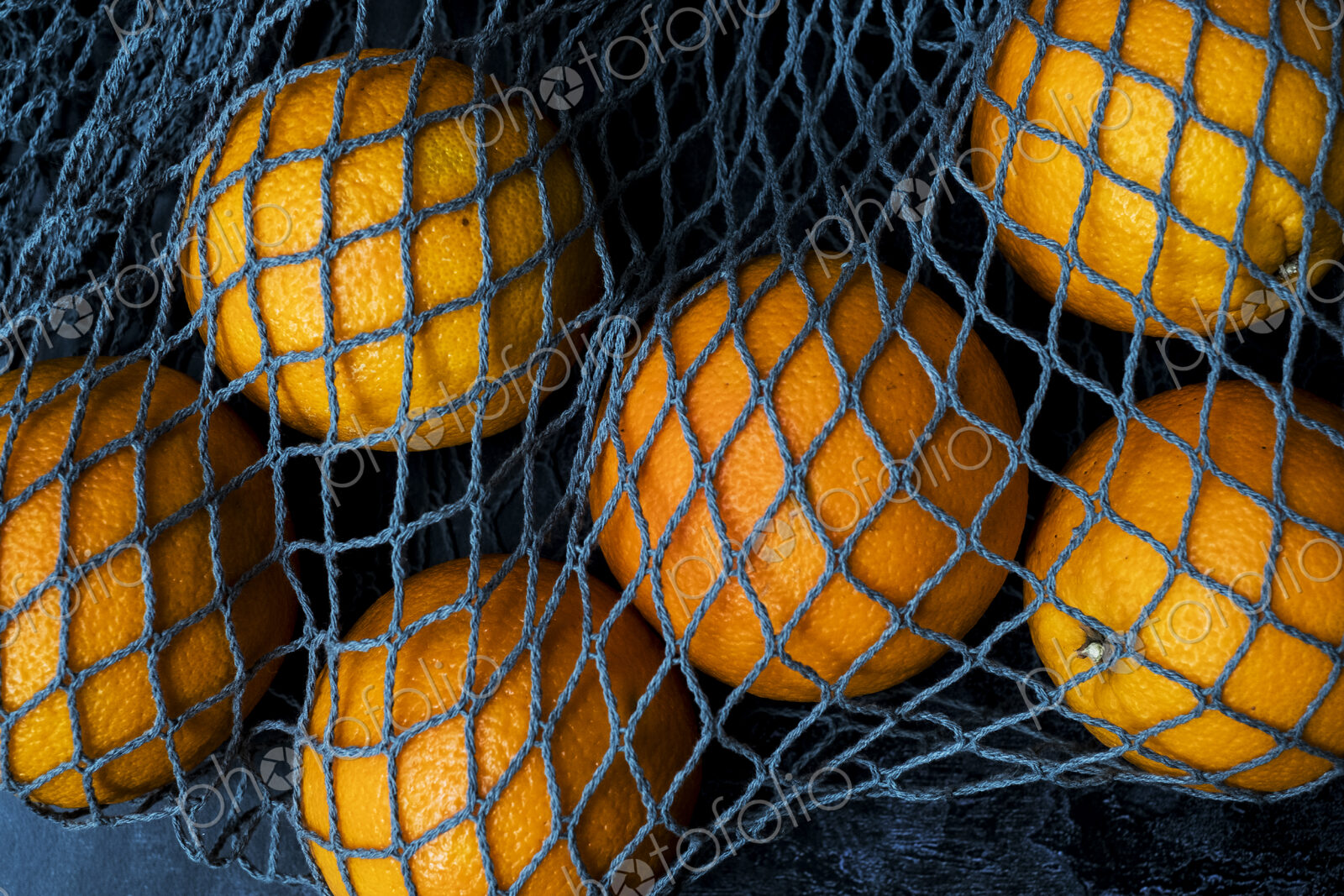 High angle close up of oranges in grey net bag on black background.