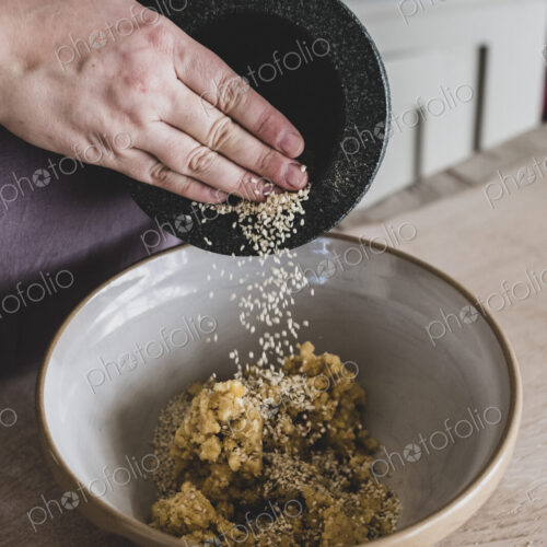 High angle close up of person adding sesame seeds to dough in a mixing bowl.