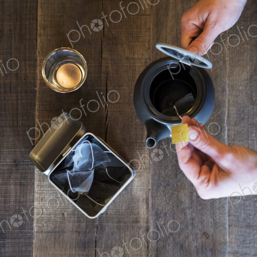 High angle close up of person adding tea bag to small black tea pot.