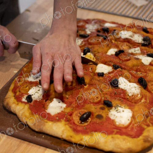 High angle close up of person cutting freshly baked pizza in an artisan bakery. High angle close up of person cutting freshly baked pizza in an artisan bakery.