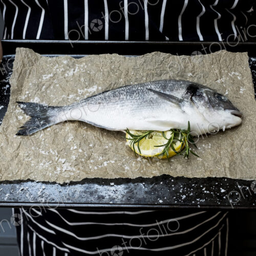 High angle close up of person holding baking tray with fresh sea bream, slices of… High angle close up of person holding baking tray with fresh sea bream, slices of…