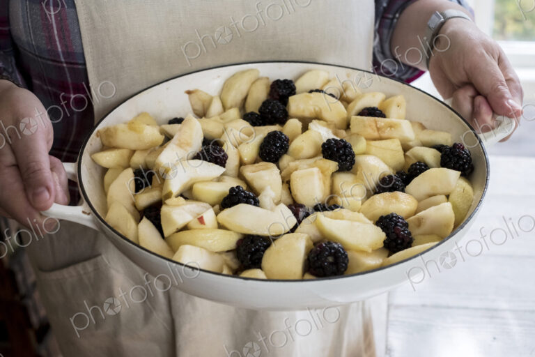 High angle close up of person holding round baking tin with apples and blackberries.