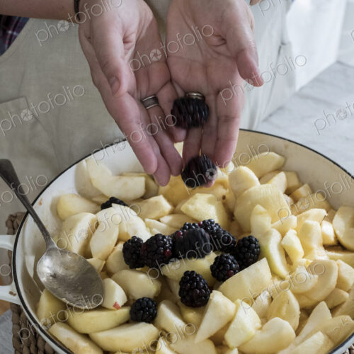 High angle close up of person placing apples and blackberries in a round baking tin.