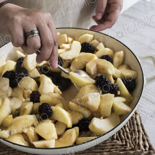 High angle close up of person placing apples and blackberries in a round baking tin.