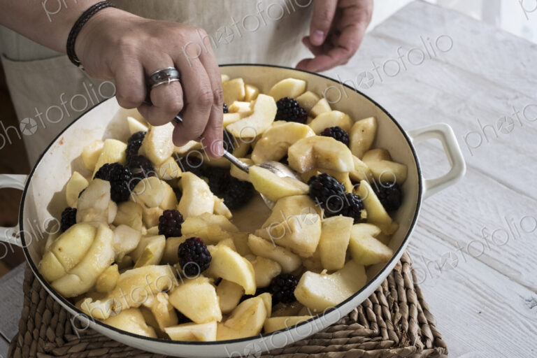 High angle close up of person placing apples and blackberries in a round baking tin.