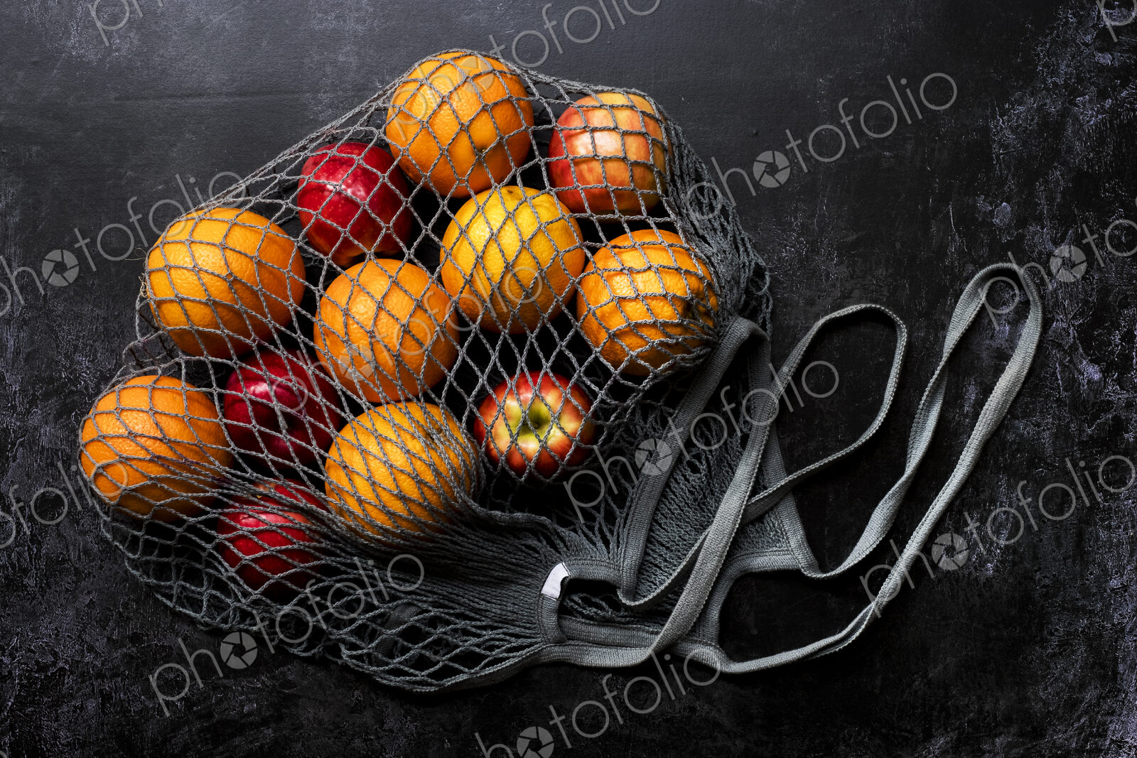 High angle close up of red apples and oranges in grey net bag on black background.