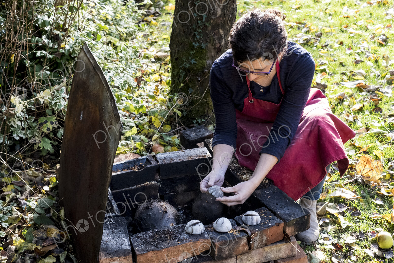 High angle close up of woman wearing red apron kneeling next to outdoor smoke fire…