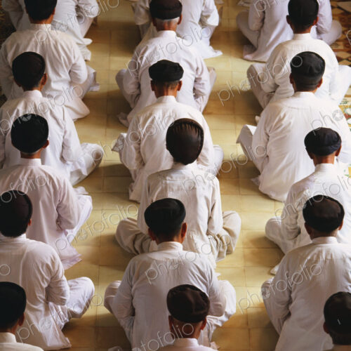 High angle rear view of of men wearing white robes sitting in rows on floor, praying.