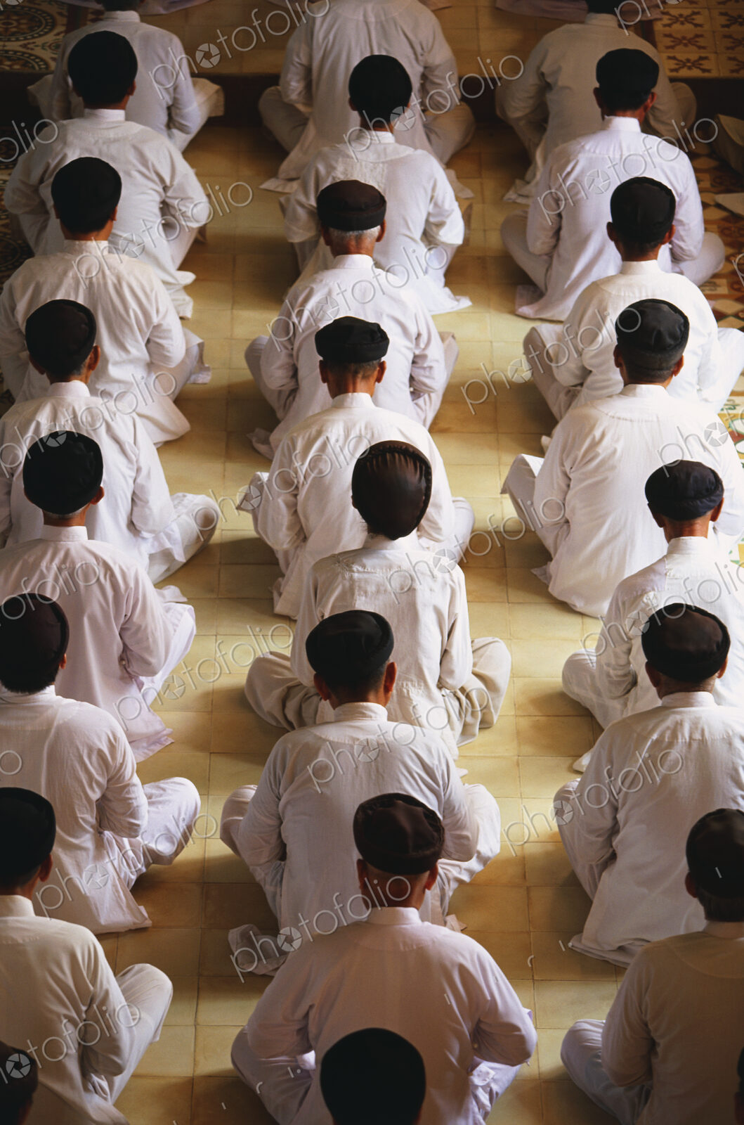 High angle rear view of of men wearing white robes sitting in rows on floor, praying.