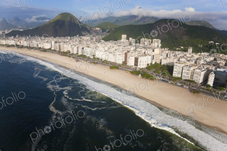 High angle view of long sandy beach lined by tall buildings, mountains in the distance.