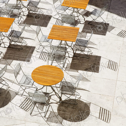 High angle view of wooden tables and metal folding chairs casting shadows onto brown… High angle view of wooden tables and metal folding chairs casting shadows onto brown…