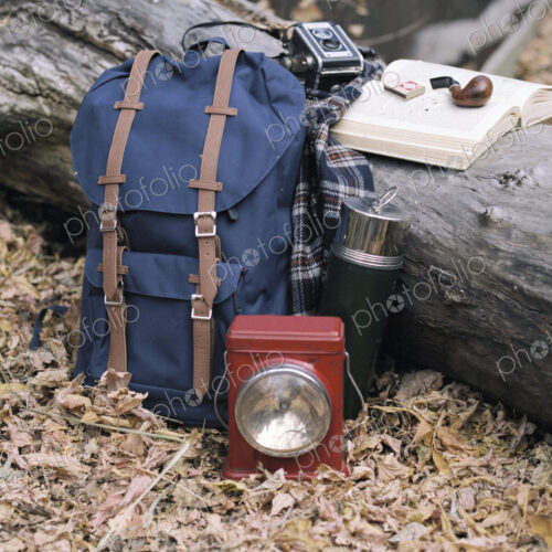 Hiking equipment, a backpack, torch, flask and camera by a tree trunk. A pipe lying on…