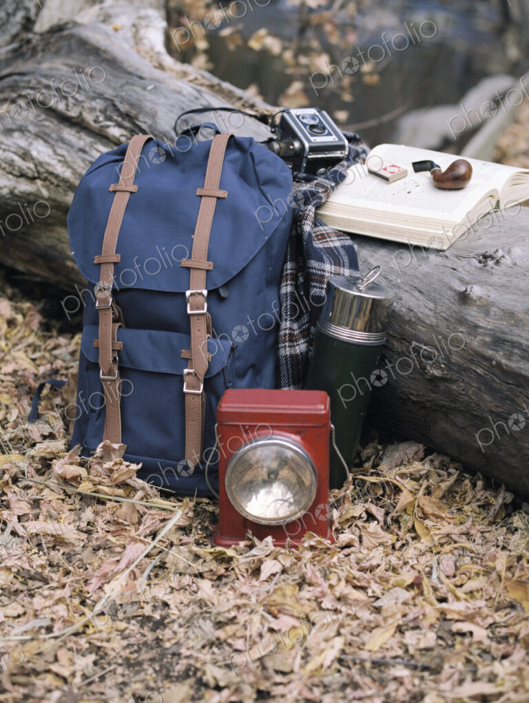 Hiking equipment, a backpack, torch, flask and camera by a tree trunk. A pipe lying on…
