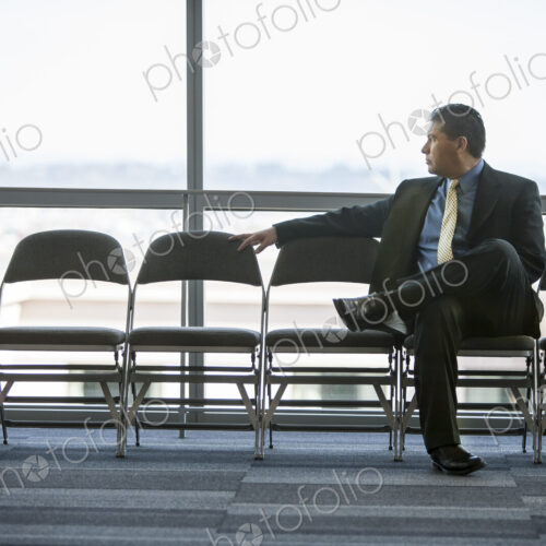 Hispanic businessman sitting in a row of chairs against a window in a conference… Hispanic businessman sitting in a row of chairs against a window in a conference…