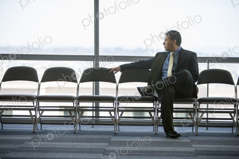 Hispanic businessman sitting in a row of chairs against a window in a conference…