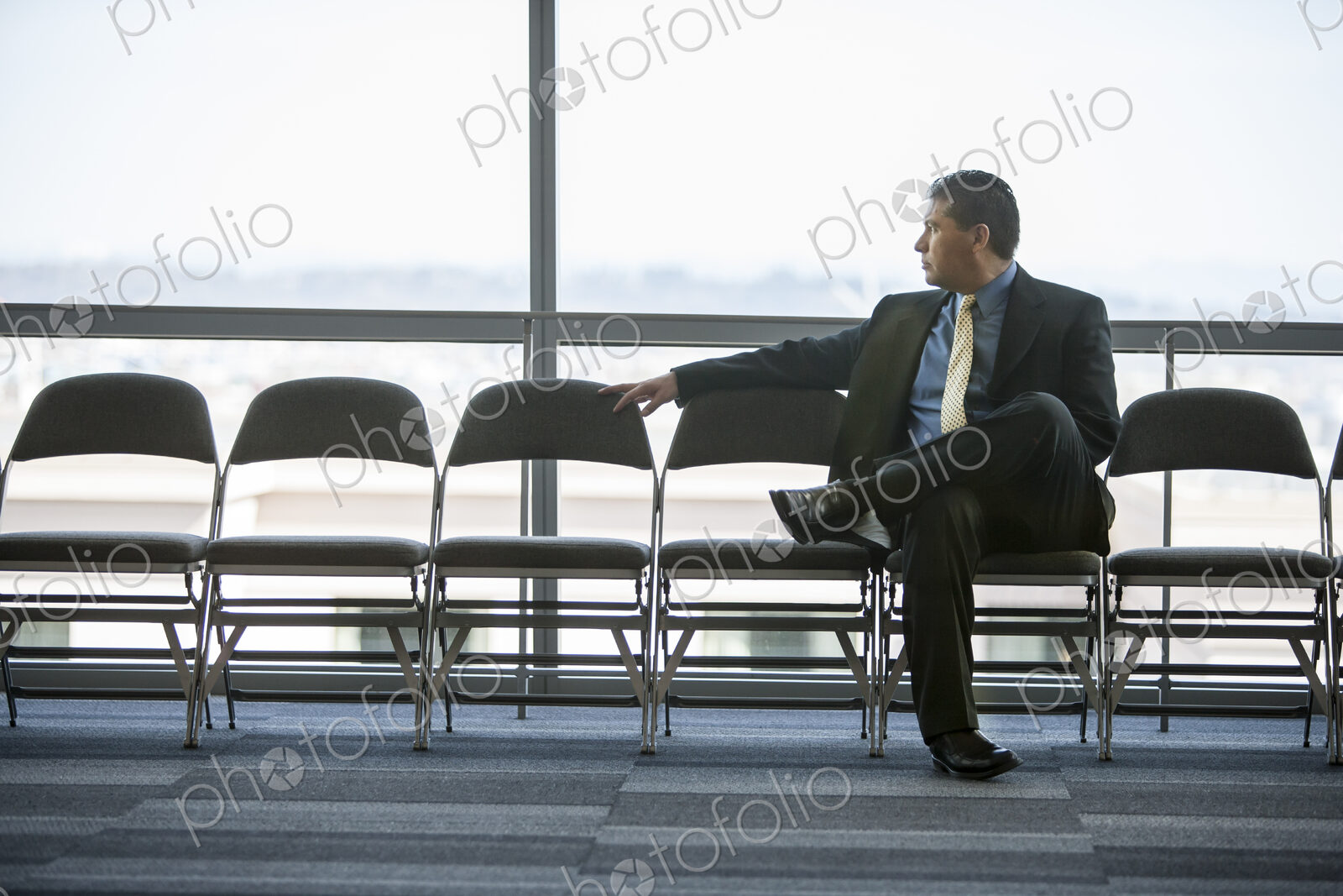 Hispanic businessman sitting in a row of chairs against a window in a conference…