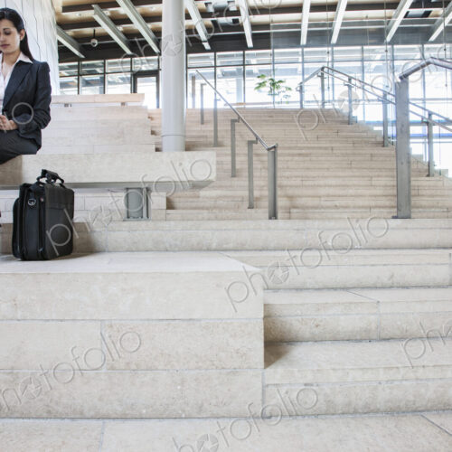 Hispanic businesswoman sitting on stairs in the lobby of a large office building.