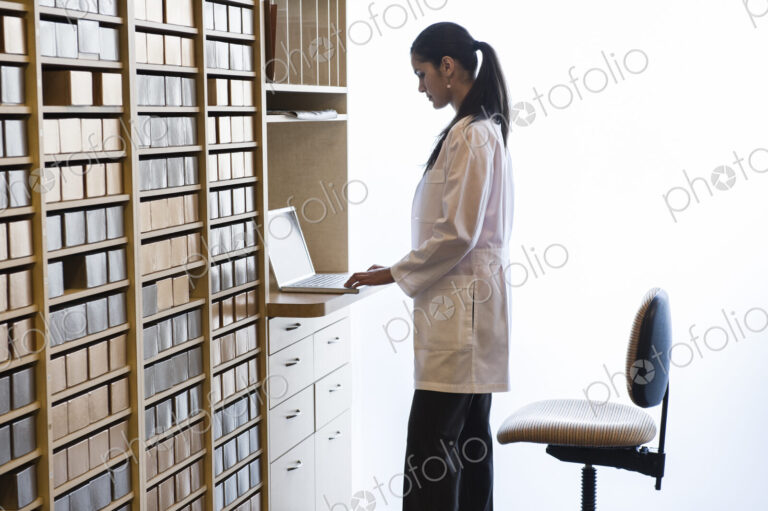 Hispanic female dentist working on a laptop computer in a cabinet file area of a…