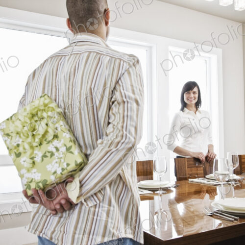 Hispanic man about to give his wife a wrapped present while standing in the dinning…