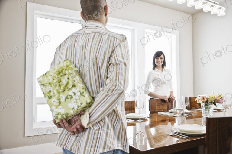 Hispanic man about to give his wife a wrapped present while standing in the dinning…