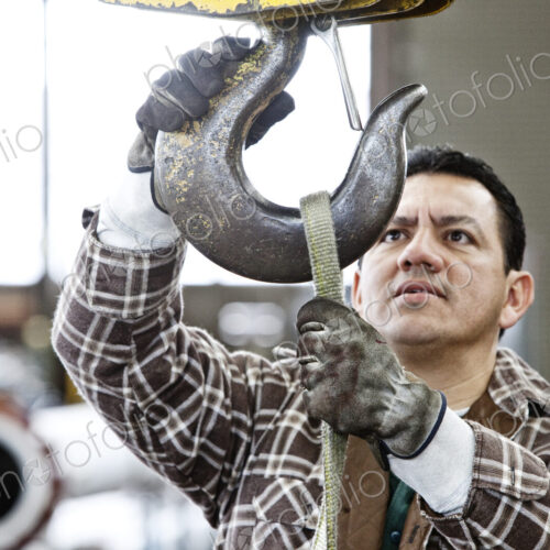 Hispanic man factory worker attaching a strap to the hook of a heavy duty overhead…