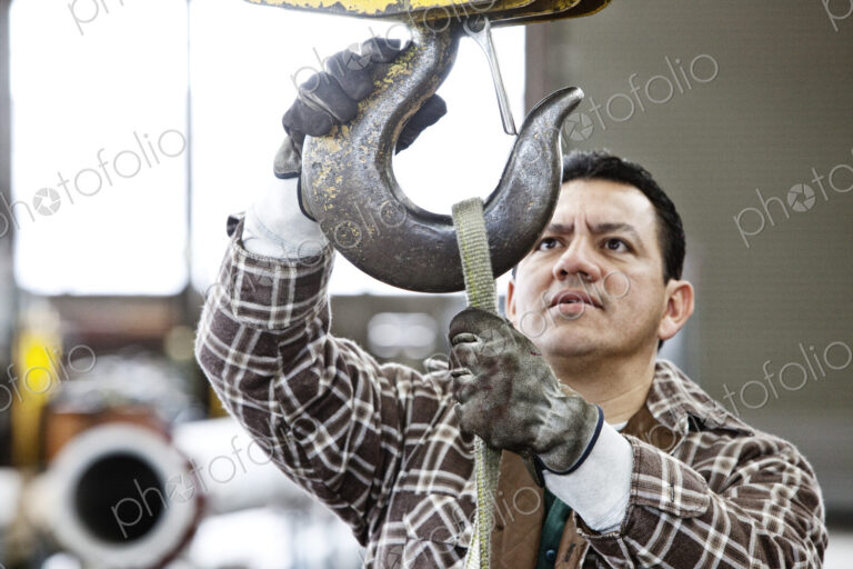 Hispanic man factory worker attaching a strap to the hook of a heavy duty overhead…