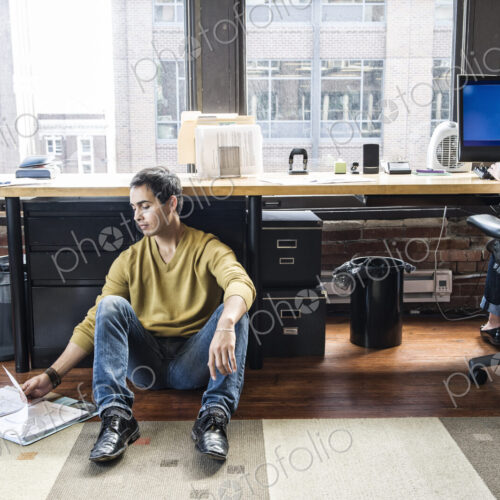 Hispanic man sitting on floor next to his work station in a creative office. Hispanic man sitting on floor next to his work station in a creative office.