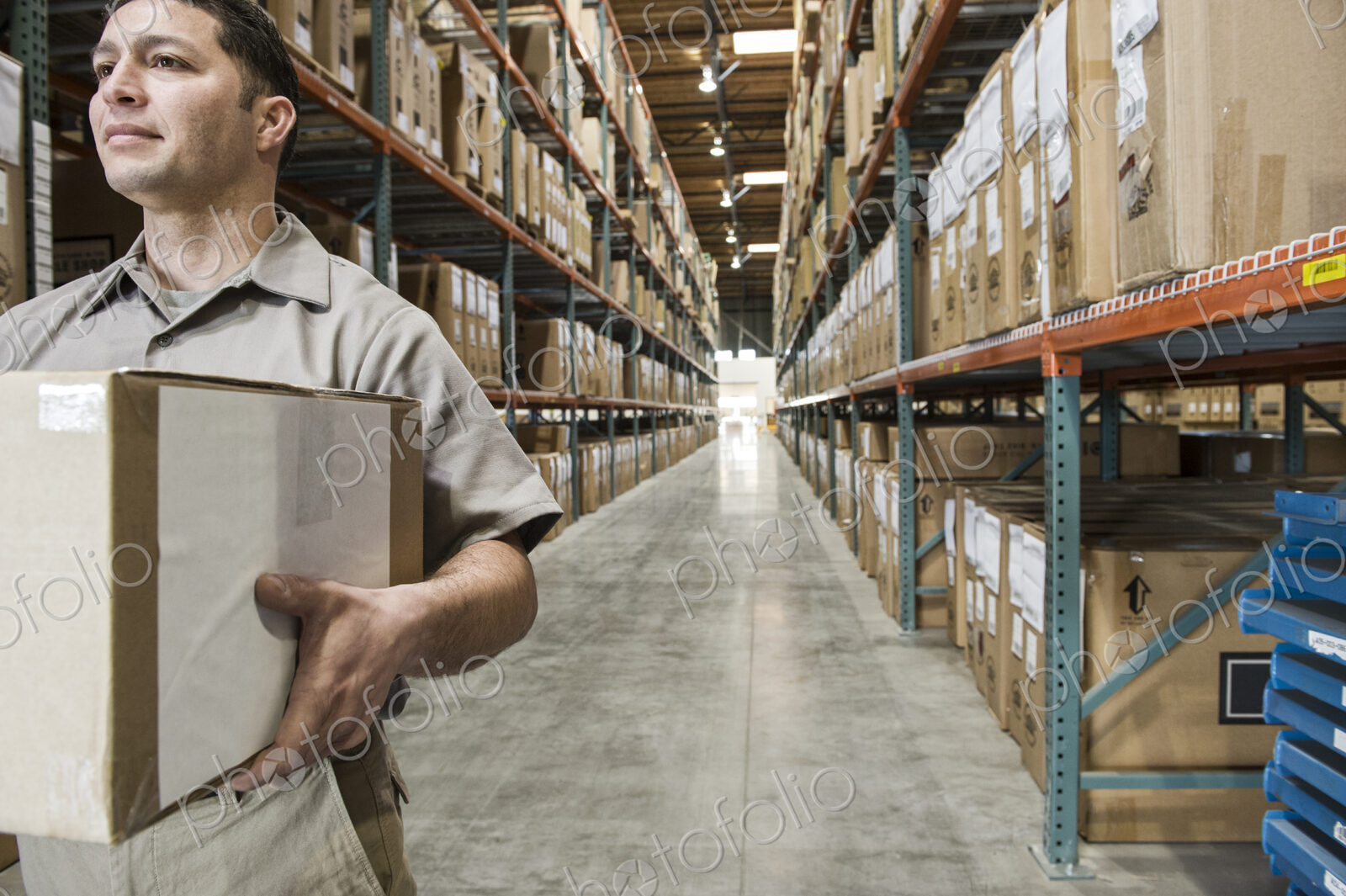 Hispanic warehouse worker carrying a box down an aisle in a large distrubiton warehouse.