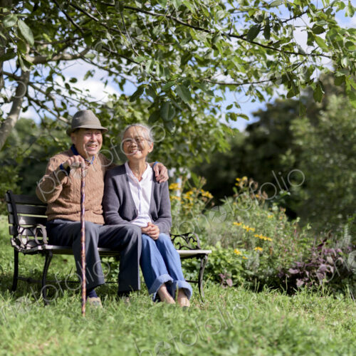 Husband and wife, elderly man wearing hat and woman sitting side by side on a bench in…