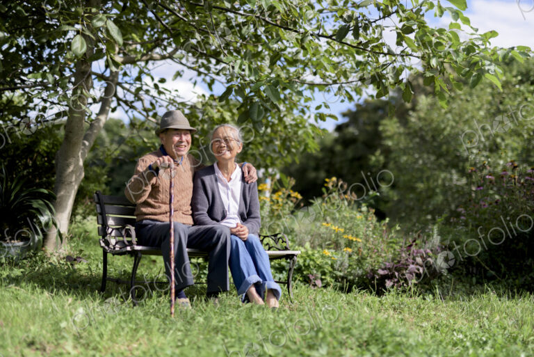Husband and wife, elderly man wearing hat and woman sitting side by side on a bench in…