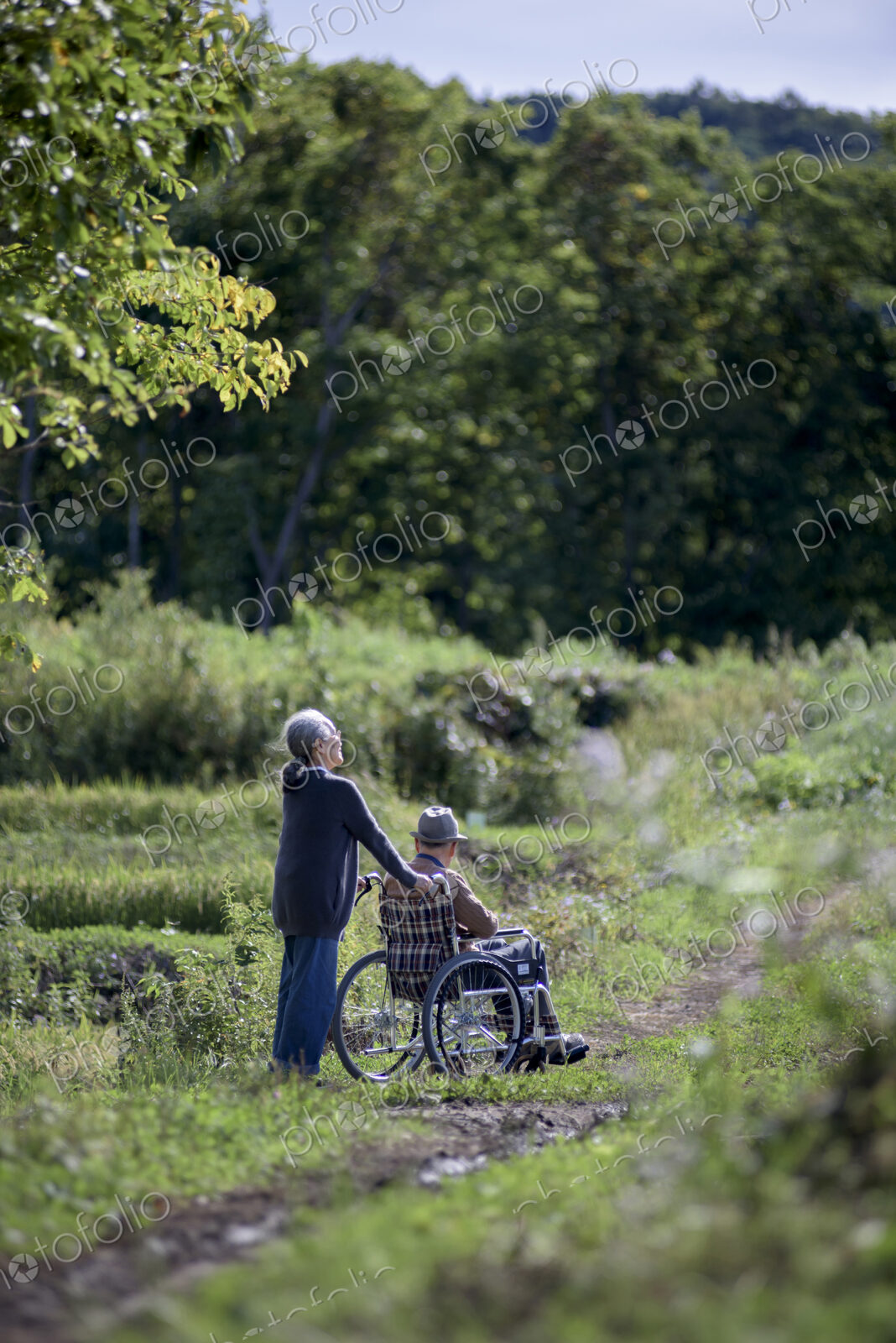 Husband and wife, elderly woman pushing elderly man wearing hat in a wheelchair.