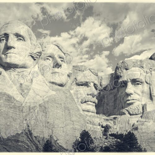 Iconic granite carvings of presidents on Mount Rushmore with clouds overhead and pines…