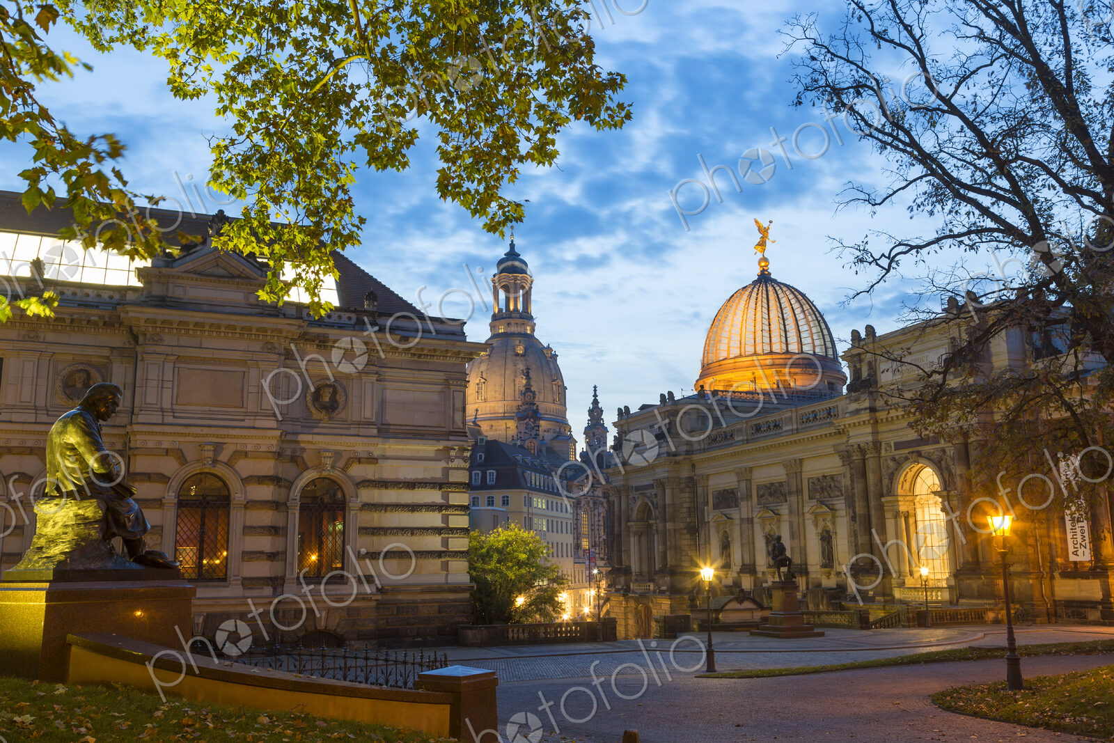 Illuminated town square with facades of domed church, college and sculptures.