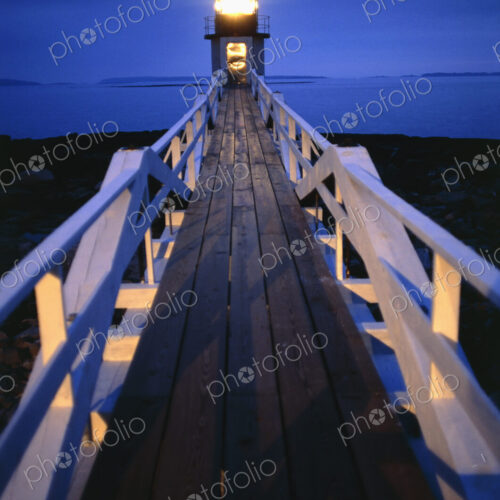 Illuminated wooden walkway towards lighthouse in the evening.
