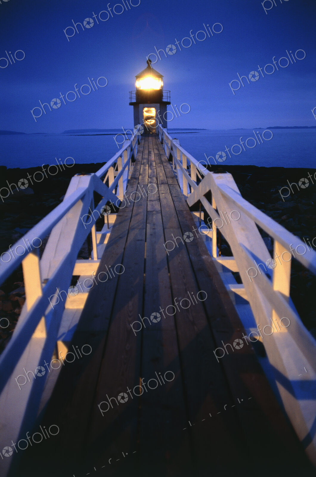 Illuminated wooden walkway towards lighthouse in the evening.