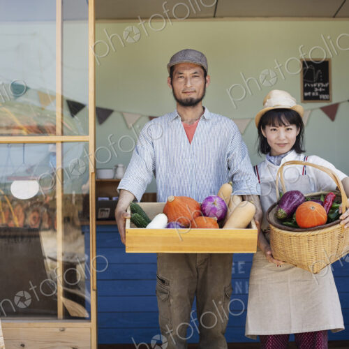 Japanese man and woman standing outside a farm shop, holding crate and basket with…