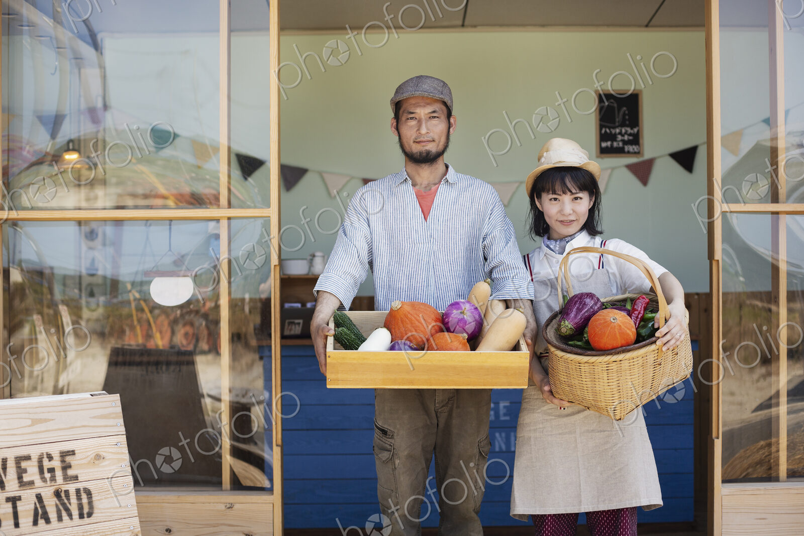 Japanese man and woman standing outside a farm shop, holding crate and basket with…