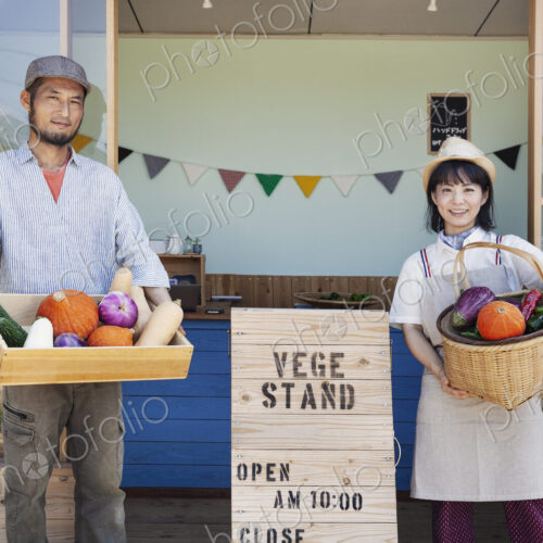 Japanese man and woman standing outside a farm shop, holding crate and basket with… Japanese man and woman standing outside a farm shop, holding crate and basket with…