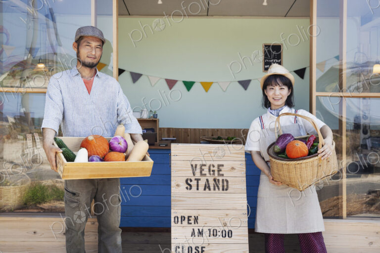Japanese man and woman standing outside a farm shop, holding crate and basket with…