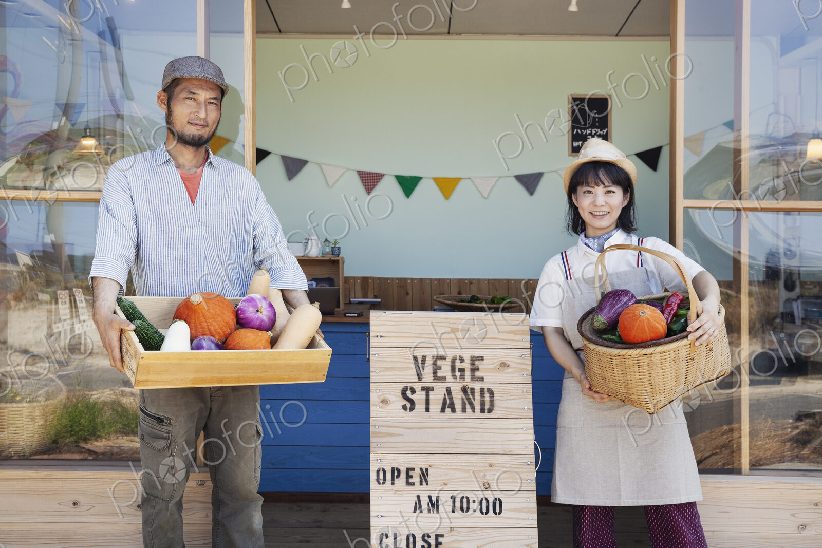 Japanese man and woman standing outside a farm shop, holding crate and basket with…