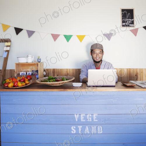 Japanese man sitting behind counter of a farm shop, working on laptop computer.