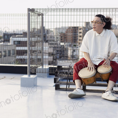 Japanese man sitting on a rooftop in an urban setting, playing drums. Japanese man sitting on a rooftop in an urban setting, playing drums.