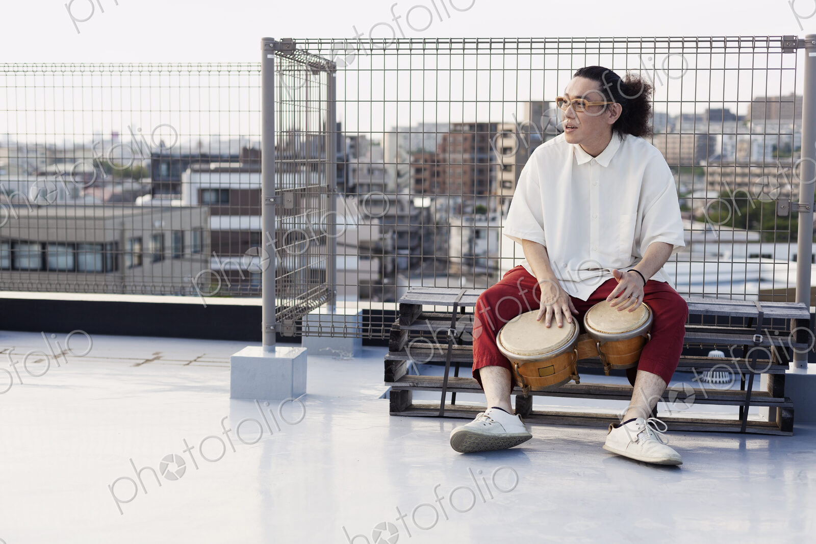Japanese man sitting on a rooftop in an urban setting, playing drums.
