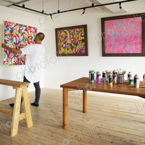 Japanese man standing in art gallery, hanging colourful painting on white wall.