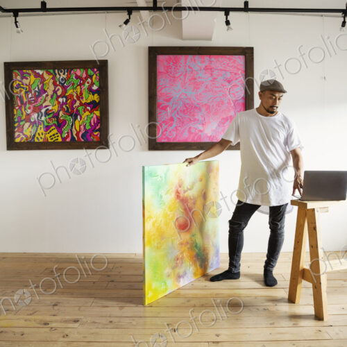 Japanese man standing in art gallery, holding abstract artwork, looking at laptop…