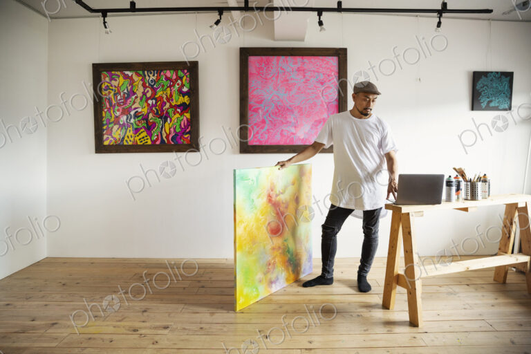 Japanese man standing in art gallery, holding abstract artwork, looking at laptop…